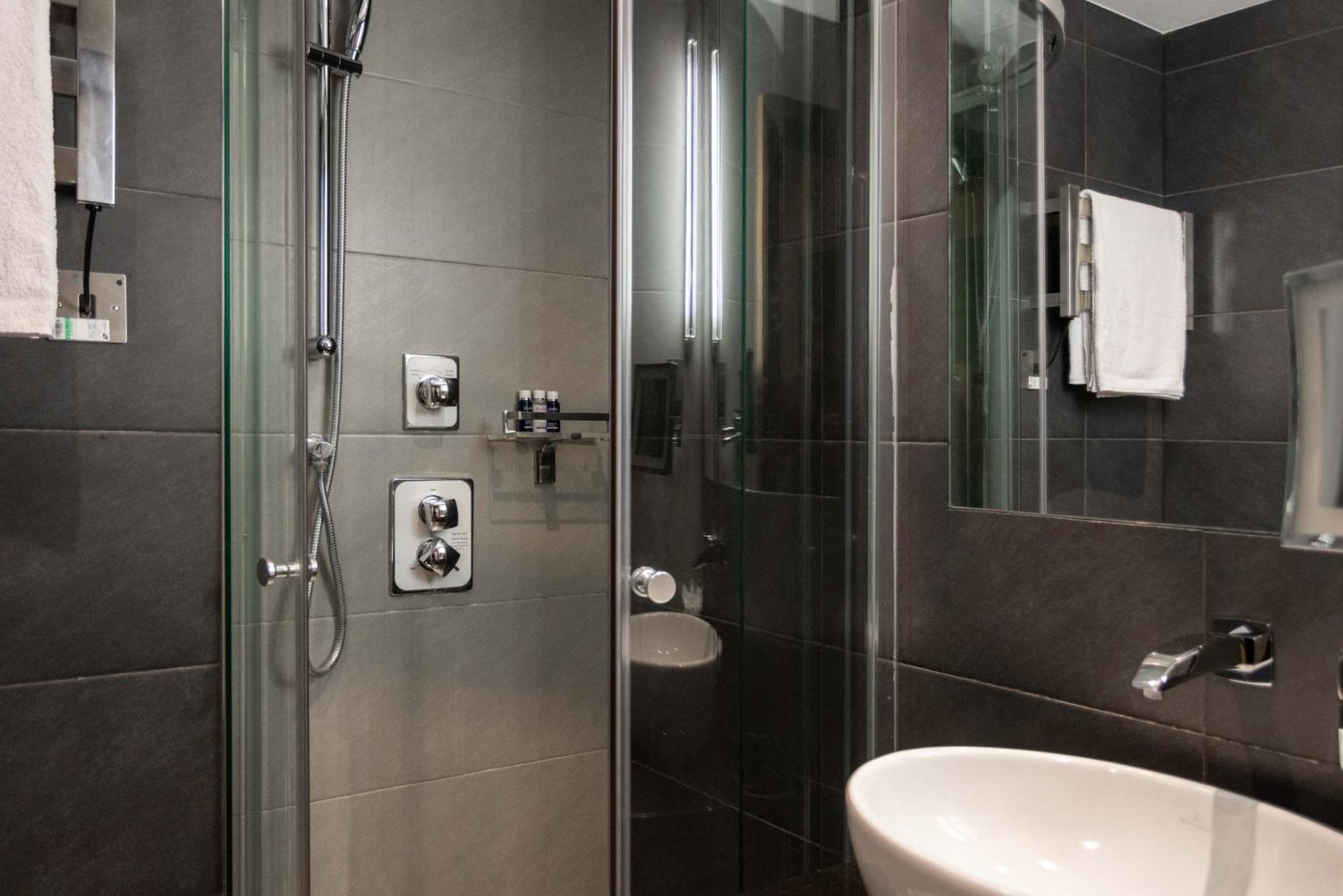 Montcalm Brewery Townhouse Hotell London Eksteriør bilde The photo shows a modern bathroom with a glass shower enclosure. The shower features a handheld showerhead and various controls mounted on the wall. The walls are tiled in dark, sleek tones, contributing to a contemporary aesthetic. There is a round 
