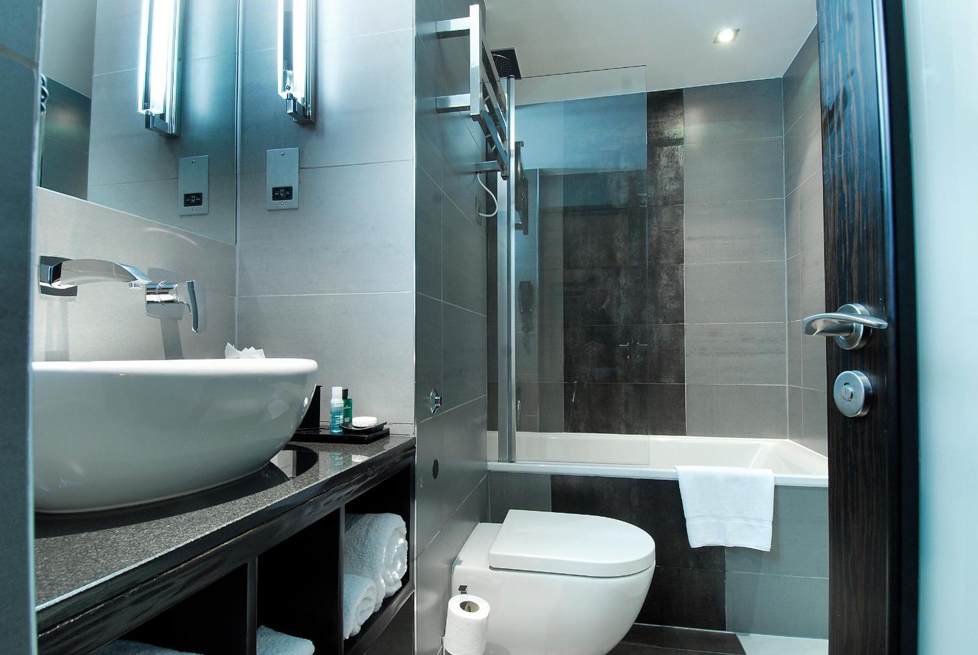 Montcalm Brewery Townhouse Hotell London Eksteriør bilde The photo shows a modern bathroom with sleek design elements. It features a curved white sink on a dark countertop, with a mirror above that is illuminated by stylish lighting fixtures. Next to the sink, there's a toilet, and on the opposite side, a 
