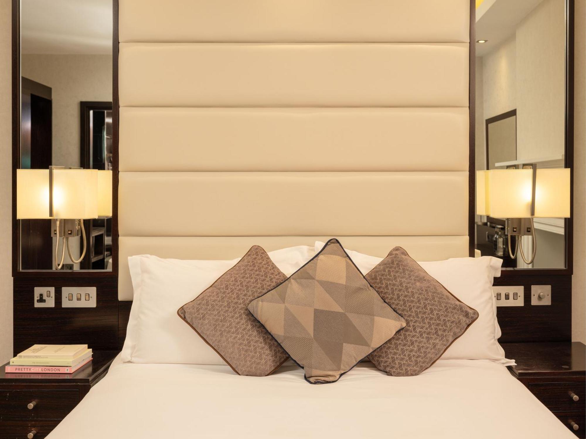 Montcalm Brewery Townhouse Hotell London Eksteriør bilde The image shows a cozy bedroom setting. The bed features a headboard upholstered in a light beige fabric. It is adorned with three decorative pillows in varying patterns and colors, placed on white bedding. On either side of the bed, there are modern