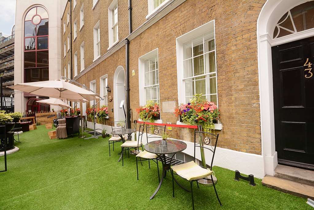 Montcalm Brewery Townhouse Hotell London Eksteriør bilde The photo depicts a charming outdoor area in front of a building with a warm, textured brick facade. There is a neatly maintained lawn, likely made of artificial grass, and several small round tables with chairs are arranged outside. Flower pots with
