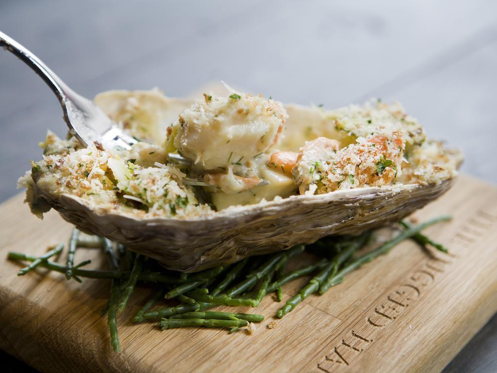 Montcalm Brewery Townhouse Hotell London Eksteriør bilde The photo shows a large oyster shell filled with a mixture that appears to include seafood, herbs, and breadcrumbs. The dish is presented on a wooden board with some greenery, possibly a type of seaweed or garnishing. A fork is placed beside the oyst