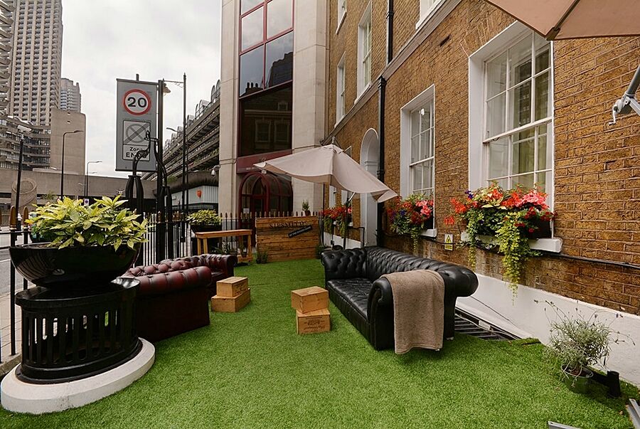 Montcalm Brewery Townhouse Hotell London Eksteriør bilde The photo depicts a charming outdoor space, likely a patio or garden area. It features two comfortable-looking leather sofas, one of which is a deep burgundy color, placed on a green artificial lawn. There are wooden crates that could serve as side t
