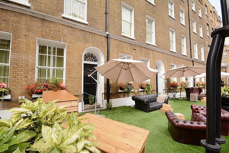Montcalm Brewery Townhouse Hotell London Eksteriør bilde The photo depicts an outdoor area of a building with a brick facade. There are several large windows visible, along with a wooden door. The setting features outdoor furniture, including couches in a dark leather finish and wooden tables. Large umbrel